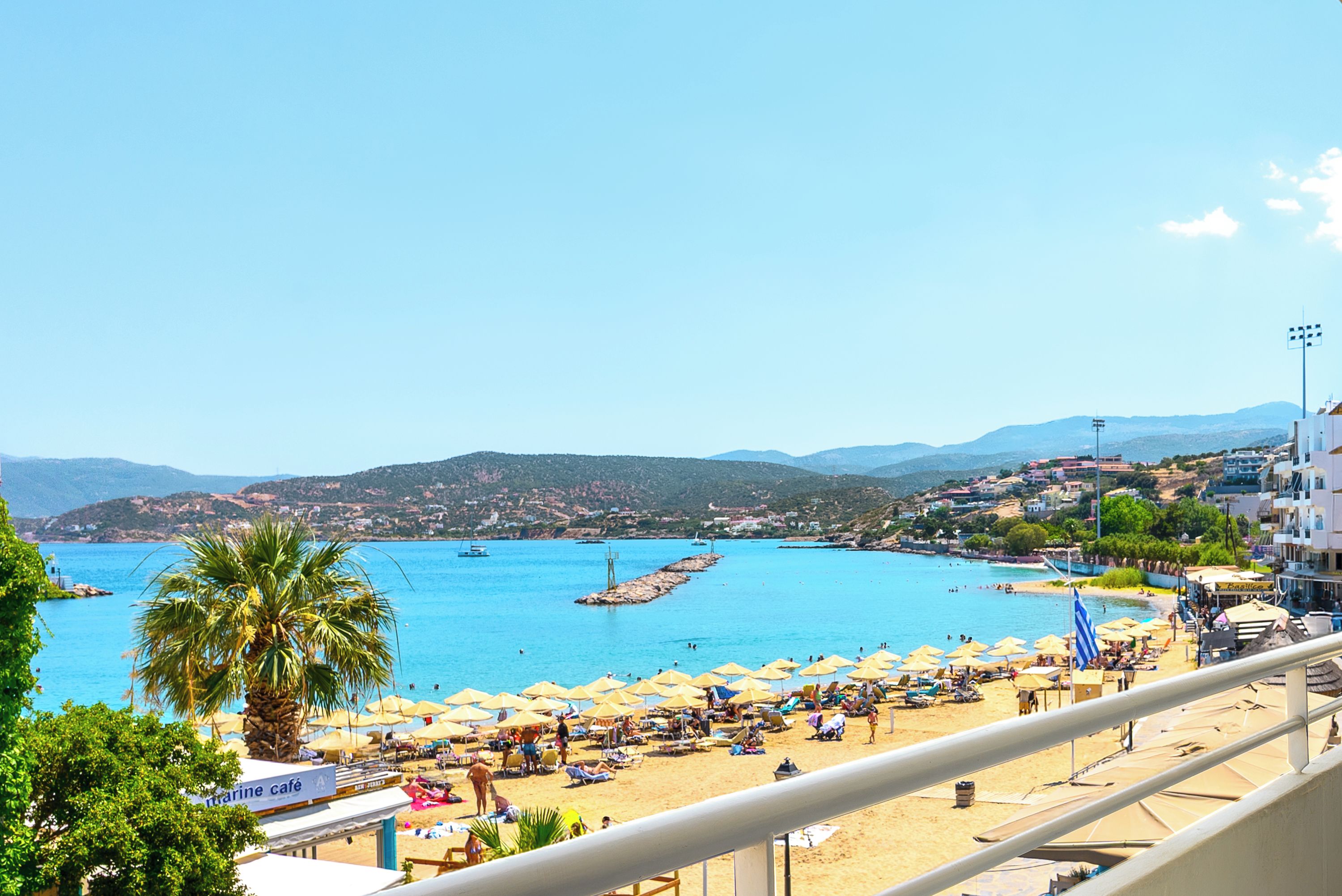 A coastal beach scene with umbrellas, sun loungers, and a clear view of the sea and distant hills.