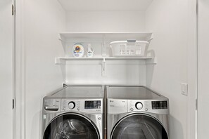 A laundry room with two front-loading washing machines and a shelf above containing laundry detergent and a laundry basket. - Modern 2BR suite with glacier views (Homer)