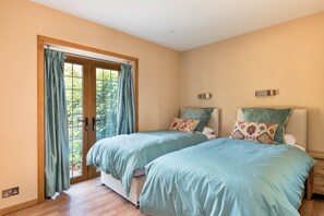 A bedroom with two beds, a wooden floor, a window with curtains, and a wall-mounted light fixture. - Riverbank Lodge Annexe (Datchet)