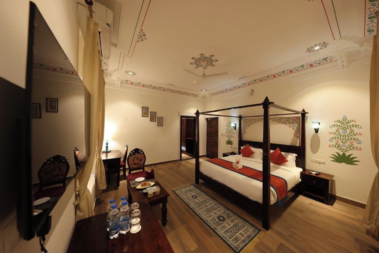 A four-poster bed with a red and white bedspread, a wooden nightstand, a table with water bottles, and a patterned rug.