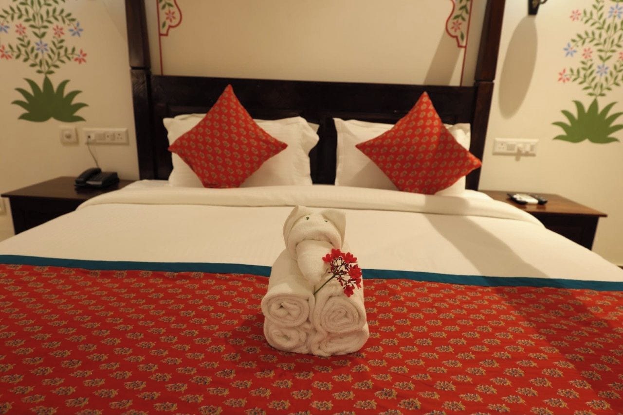 A hotel room with a neatly made bed, red patterned bedspread, and decorative pillows.