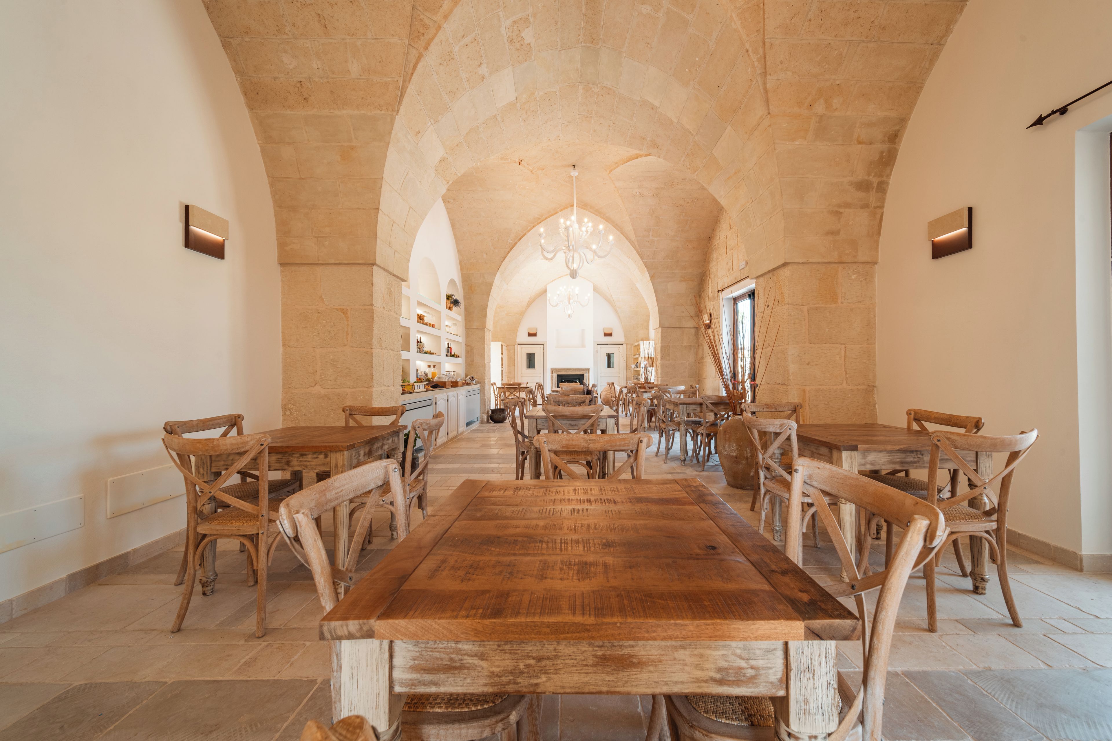 A spacious dining hall with a long wooden table and chairs, arched stone ceiling, and a chandelier.