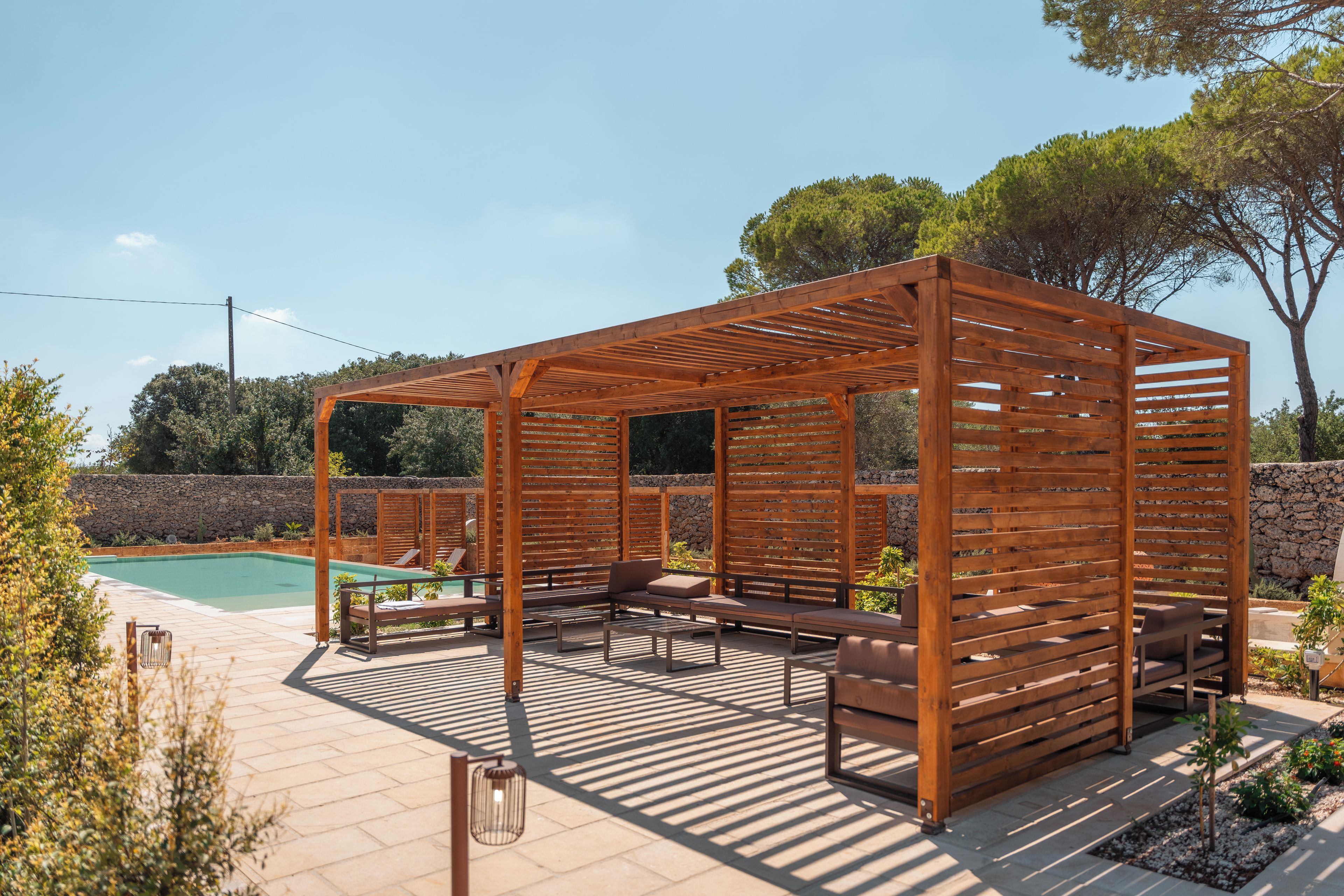 Una estructura de pérgola de madera con asientos y una piscina al fondo.