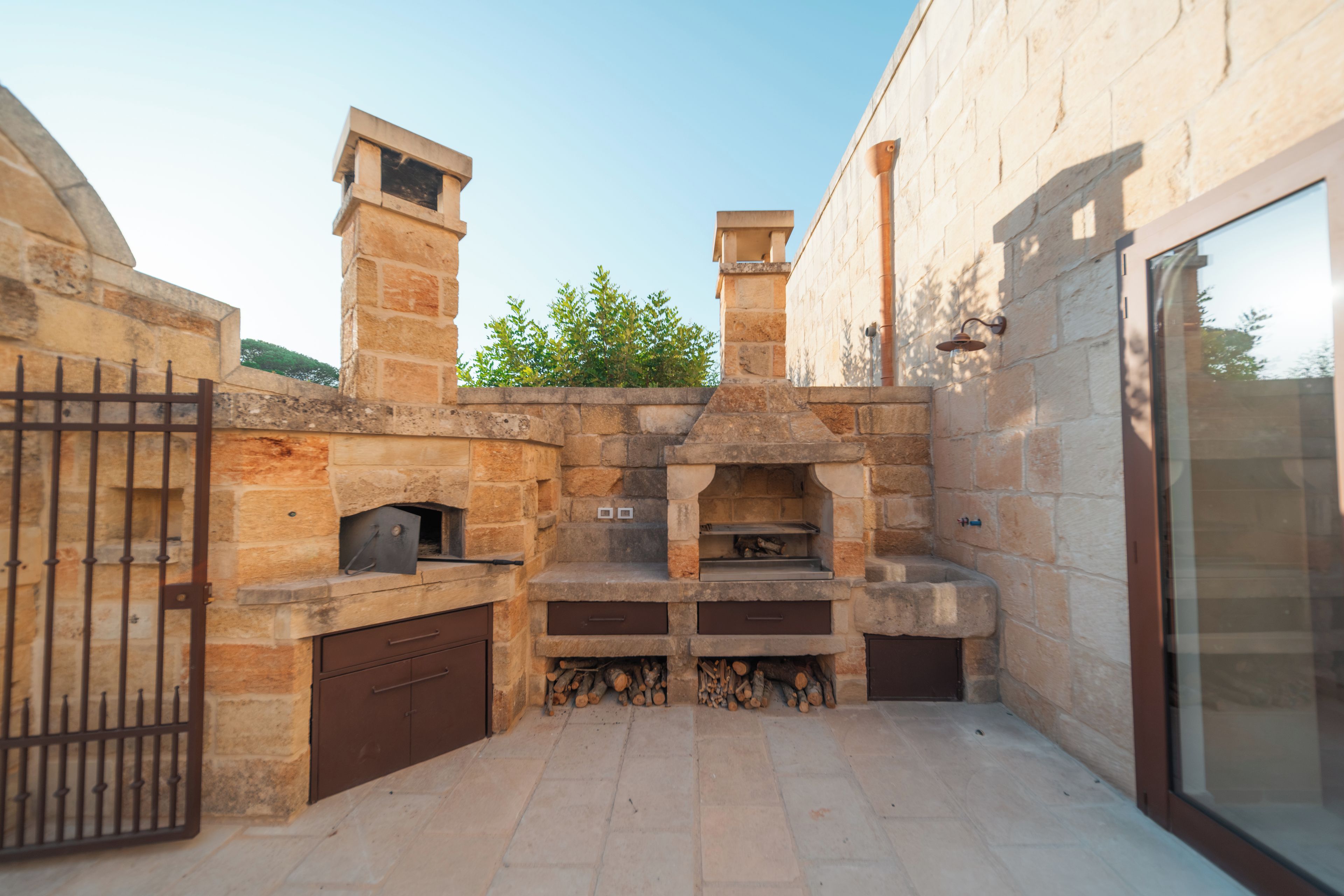 An outdoor stone barbecue area with a chimney, a fire pit, and a metal gate.