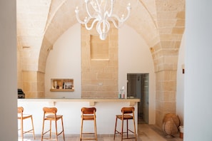 Uma área de bar com balcão branco, cadeiras de madeira e um candelabro.
