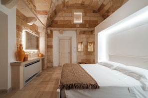 Un dormitorio con pared de piedra, cama con ropa blanca, puerta de madera y una ventana pequeña.