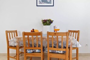 Une table à manger recouverte d’une nappe à motifs floraux, entourée de chaises en bois avec des coussins à carreaux. Un vase rempli de fleurs et une bouteille de jus d’orange sont posés sur la table.