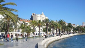 Une promenade en bord de mer, bordée de palmiers, avec une fontaine aux formes sinueuses et des gens qui se promènent.