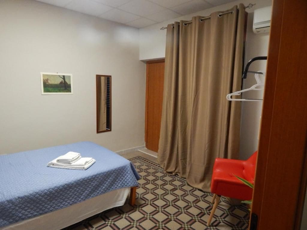 Une petite pièce comprenant un lit, une chaise, un rideau et un climatiseur fixé au mur.