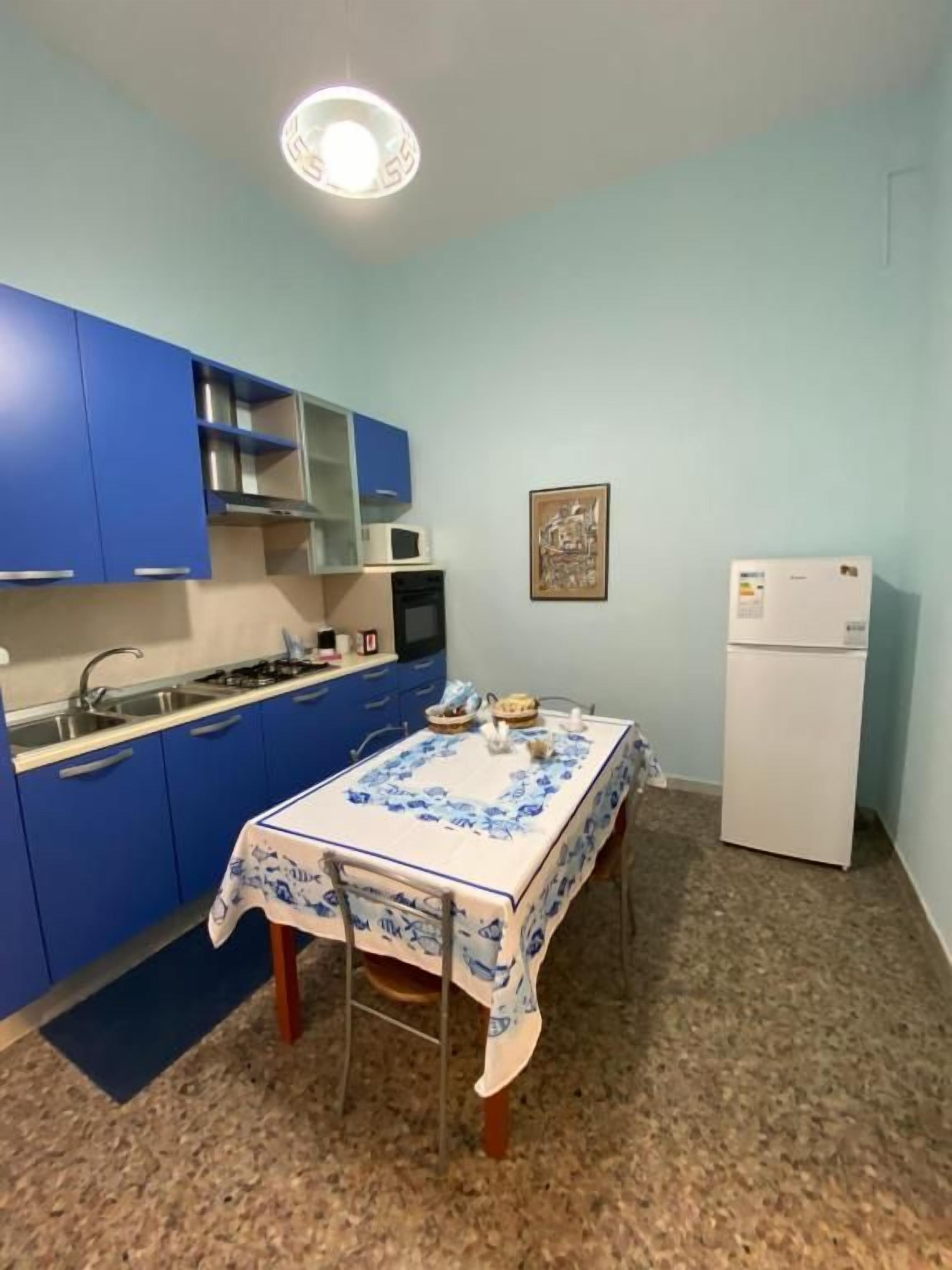 Uma cozinha compacta com armários azuis, uma geladeira branca e uma mesa com toalha de mesa florida.