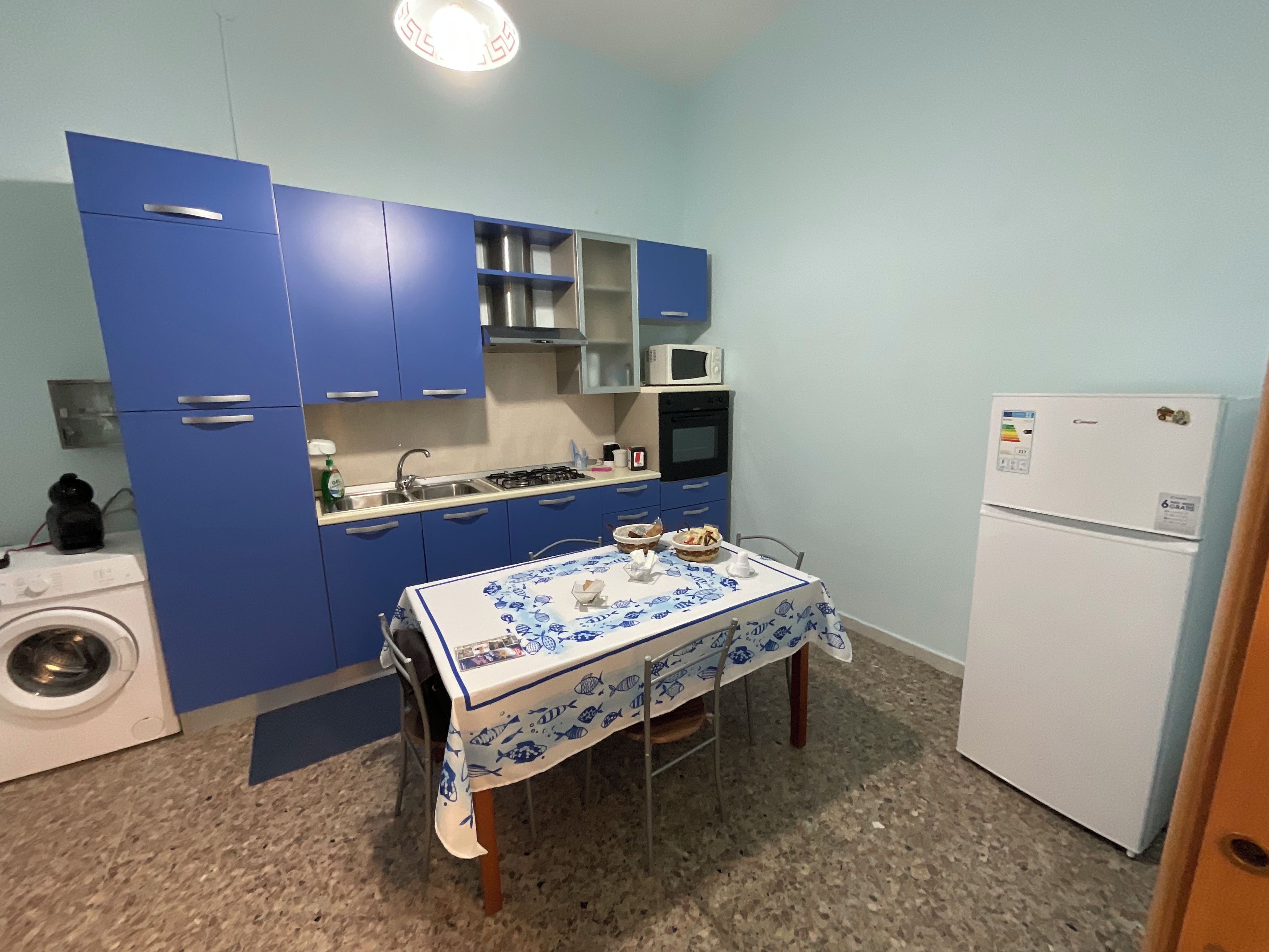 Une cuisine de taille réduite avec des meubles bleus, une table recouverte d’une nappe à motifs de poissons, un lave-linge et un réfrigérateur.
