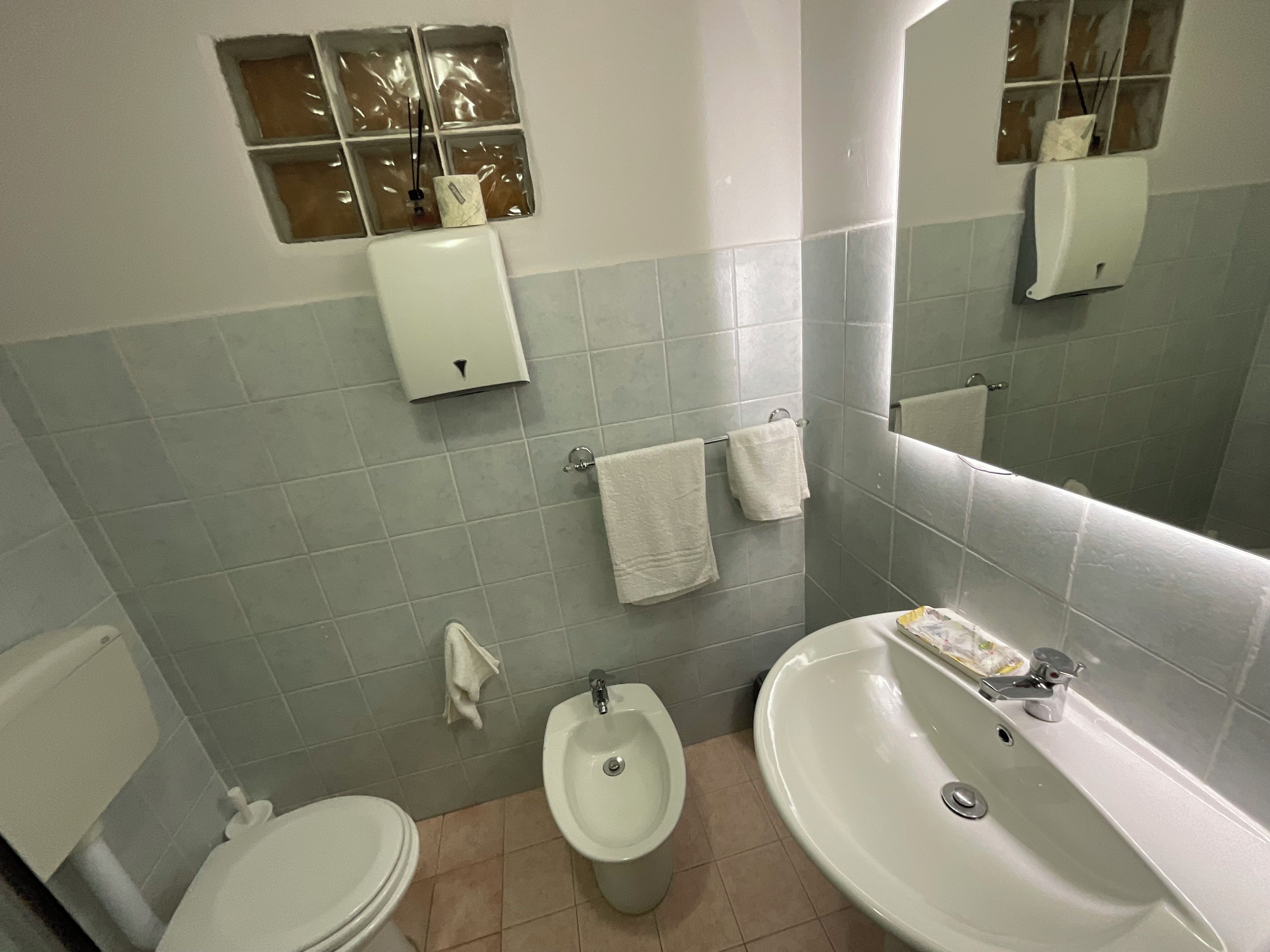 Een badkamer met toilet, bidet en wastafel. Handdoeken hangen aan de muur en er is een zeepdispenser.