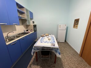 Una cocina compacta con armarios azules, un refrigerador blanco y una mesa con mantel decorado con motivos de peces.