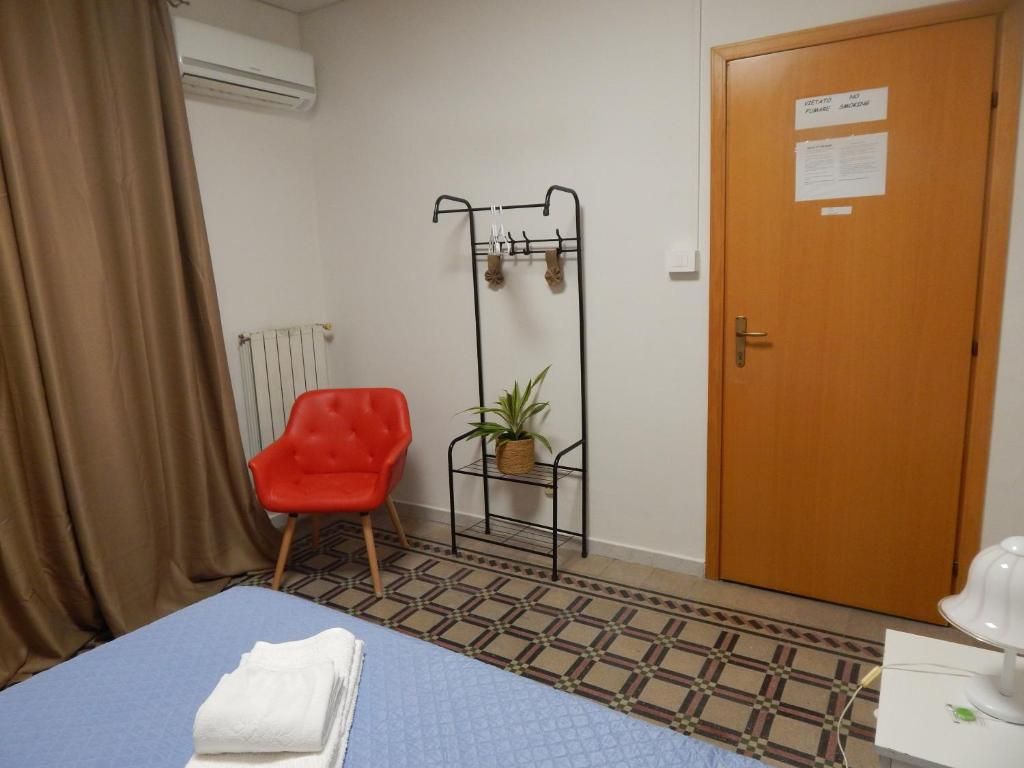Una habitación con una cama, una silla roja, un radiador, una puerta con un aviso y una planta sobre un soporte metálico.