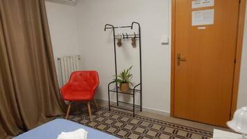 Una habitación con una cama, una silla roja, un radiador, una puerta con un aviso y una planta sobre un soporte metálico.