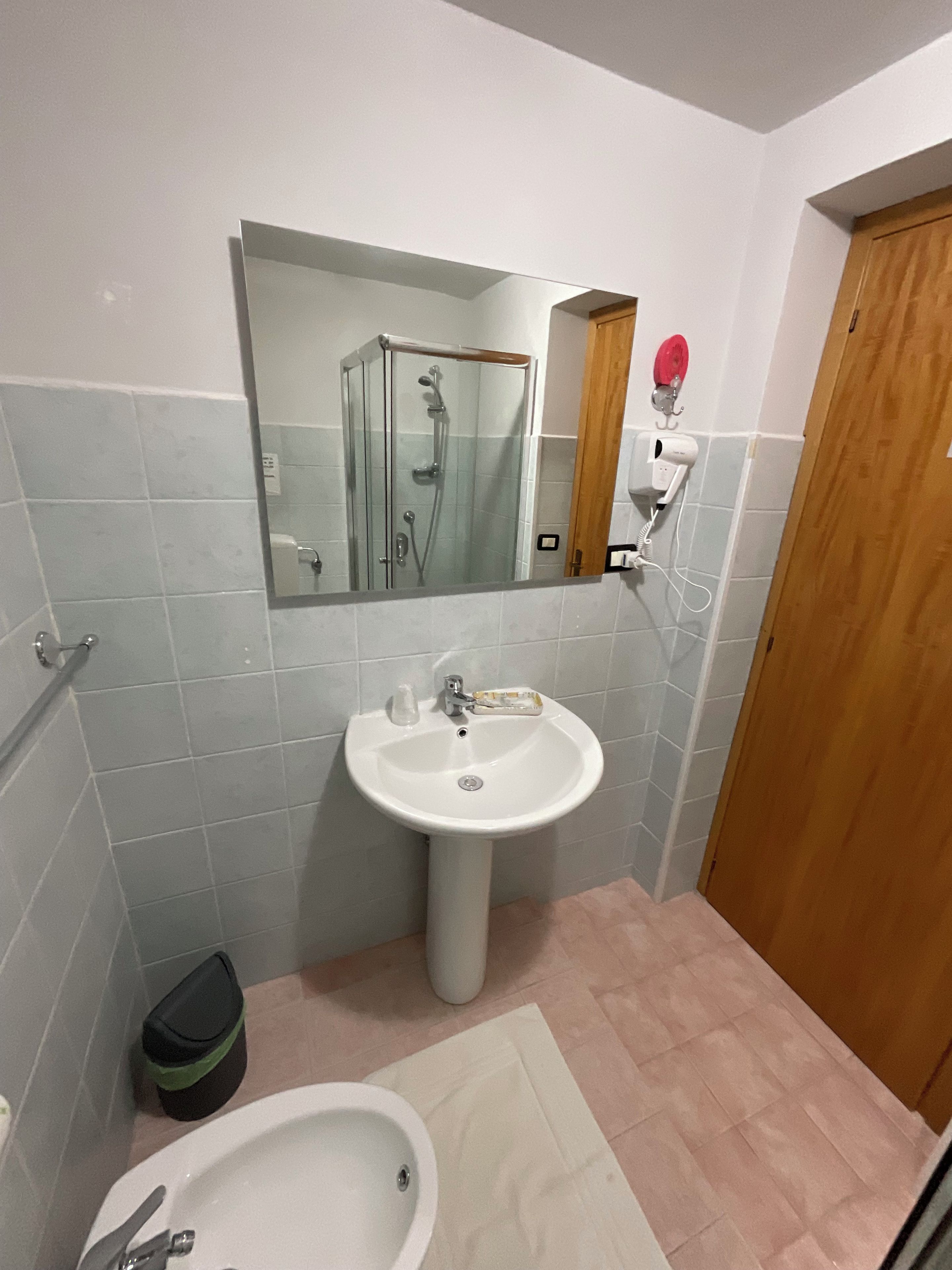 Ein Badezimmer mit einem Standwaschbecken, einem Spiegel darüber, einer Duschkabine und einer Holztür.
