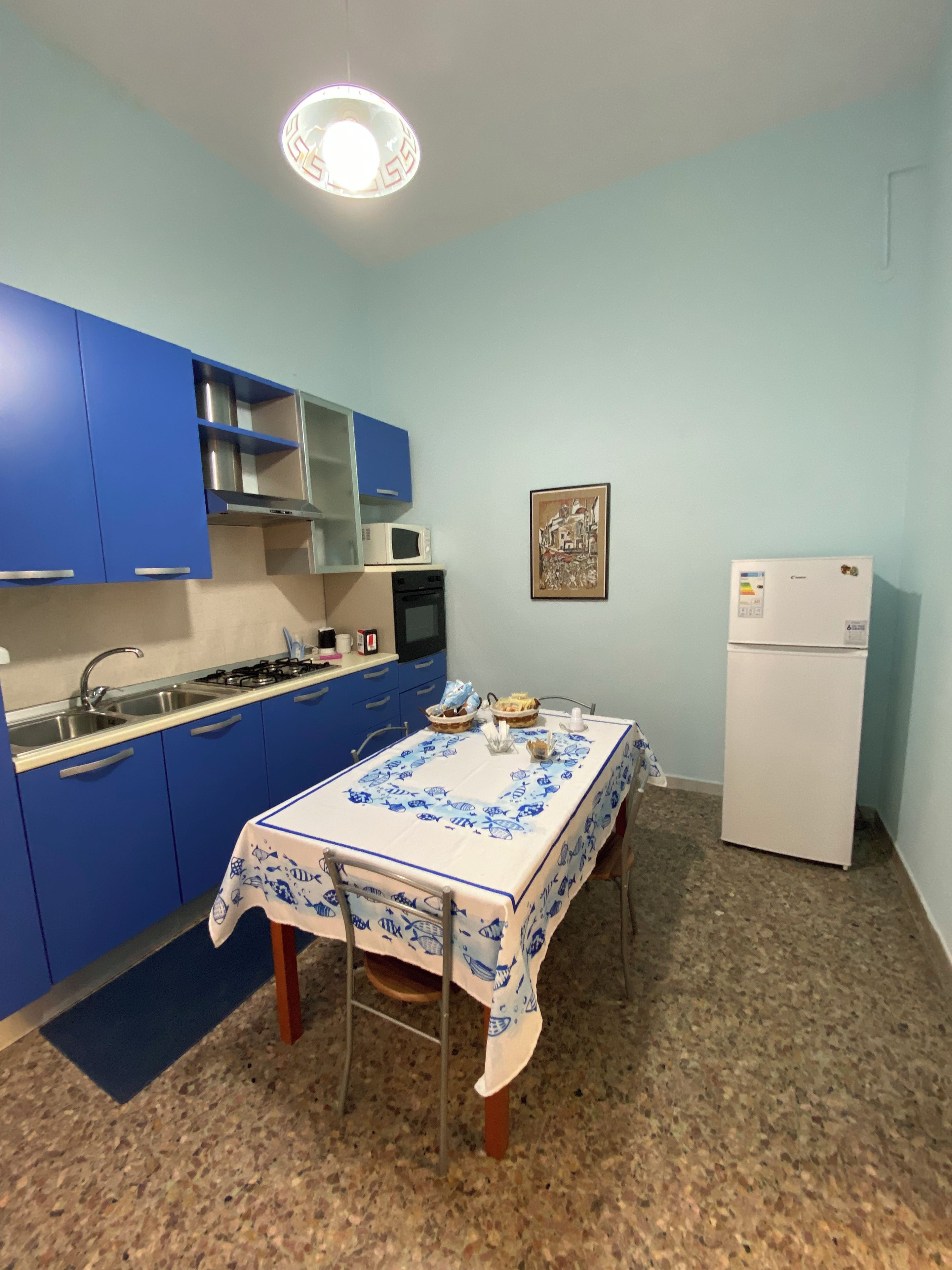 Une cuisine de taille réduite avec des meubles bleus, un réfrigérateur blanc et une table à manger recouverte d’une nappe à motifs de poissons.