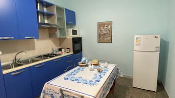 Une cuisine de taille réduite avec des meubles bleus, un réfrigérateur blanc et une table à manger recouverte d’une nappe à motifs de poissons.