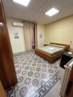 Una habitación con cama, radiador, ventana con cortinas y baldosas de suelo estampadas.