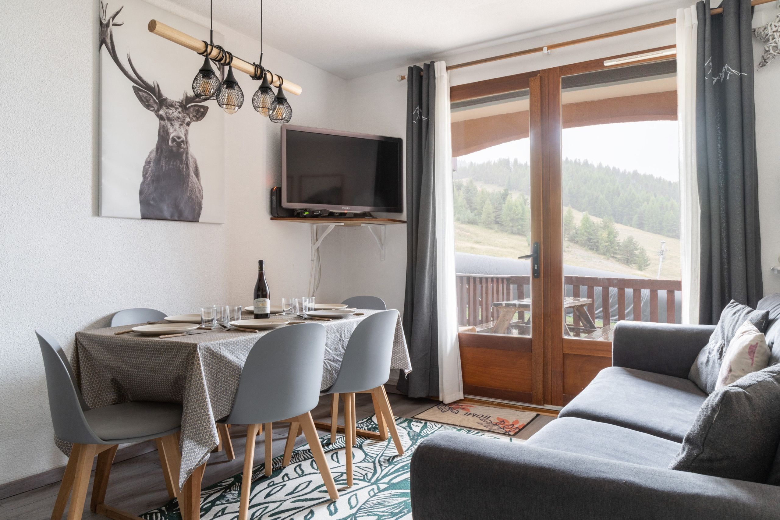 A dining area with a table set for four, a television mounted on the wall, and a view of a forested landscape through a sliding glass door.