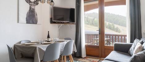 A dining area with a table set for four, a television mounted on the wall, and a view of a forested landscape through a sliding glass door.