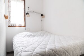 A single-bed room with a white quilted bedspread, a wall-mounted lamp, and a window with a lace curtain. - Facing the slopes, 6-person apartment with terrace (Le Dévoluy)