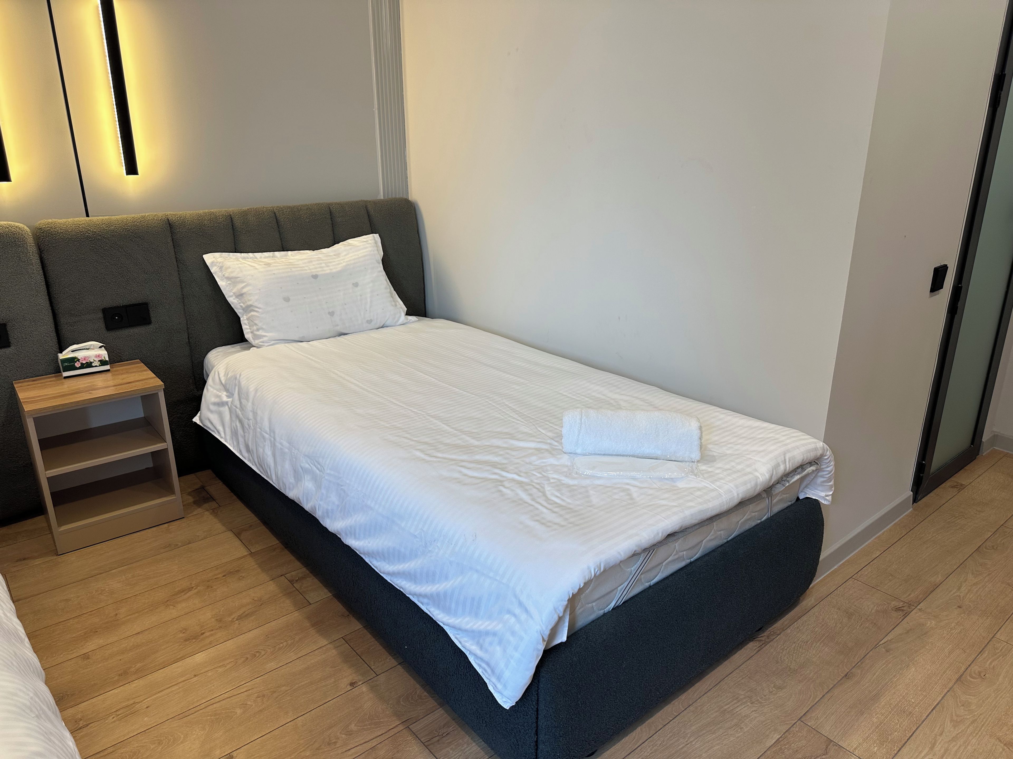 Un letto singolo con una testata grigia, biancheria da letto bianca e un comodino in legno con un telefono.