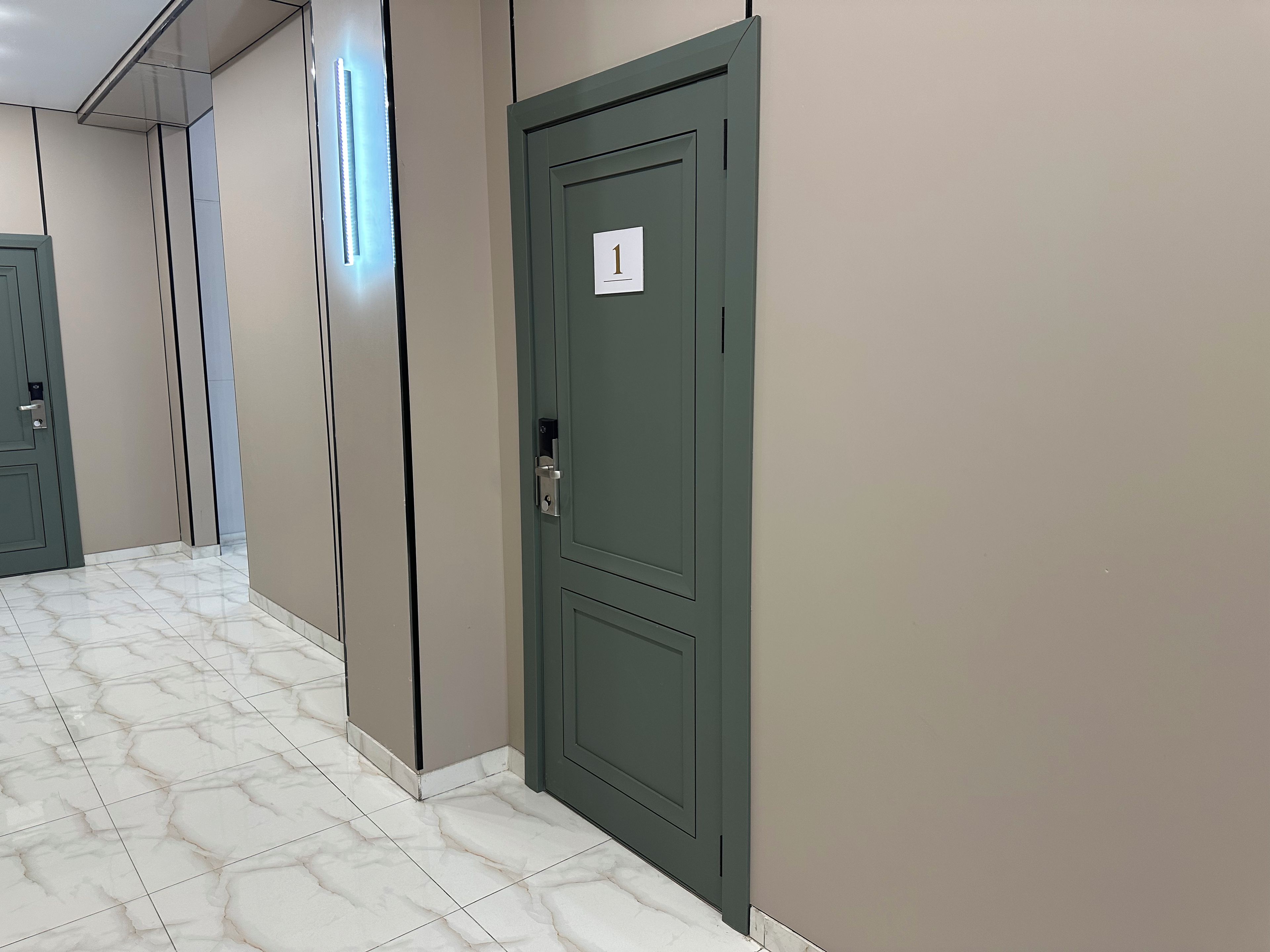 A hallway with a green door marked with the number 1, white tiled flooring, and a light fixture on the wall.