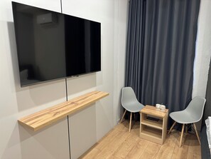 A modern room with a flat-screen TV mounted on a wall, a wooden shelf below, two grey chairs, a small table with a glass, and a dark curtain.