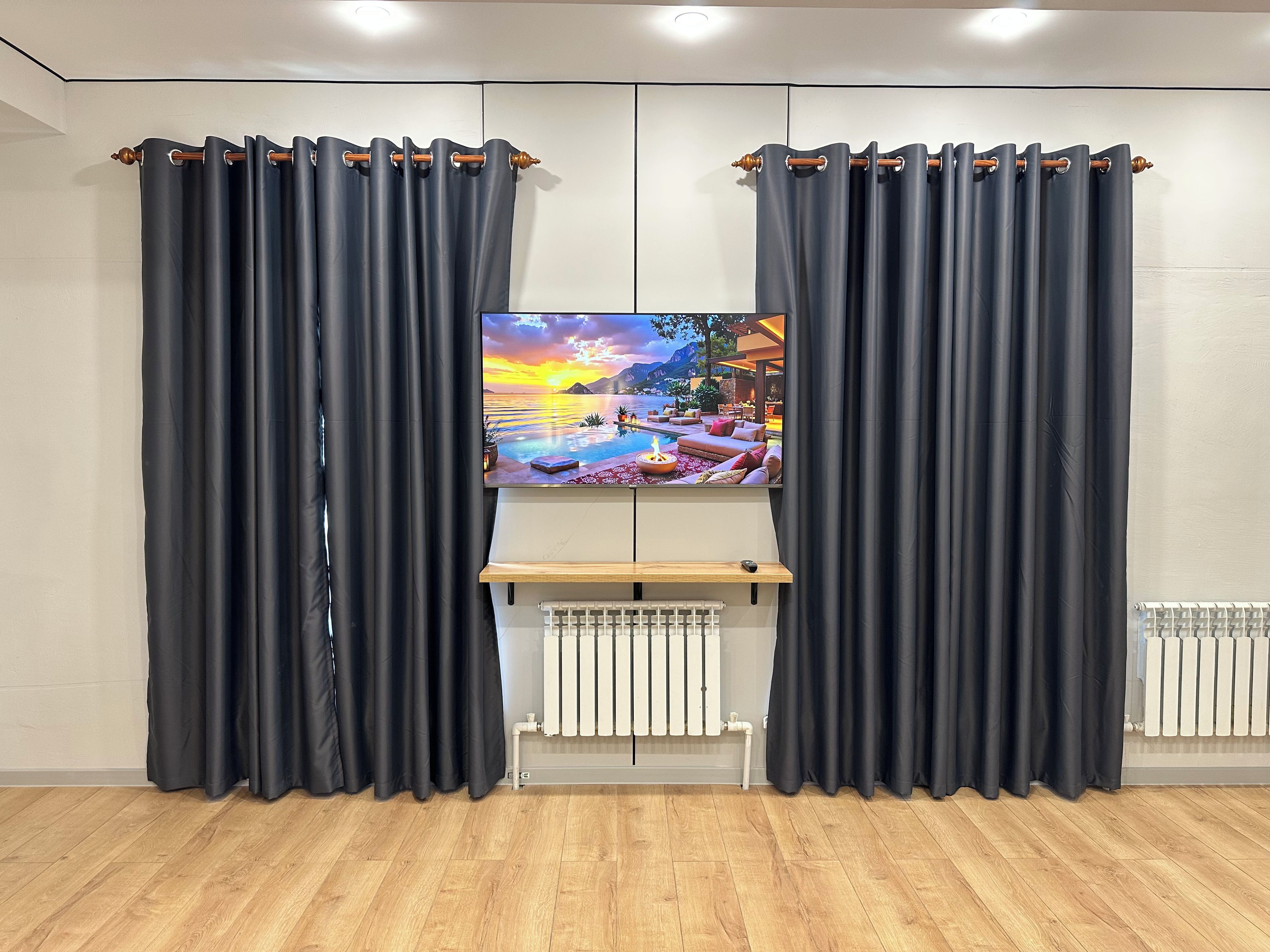 Una stanza con una TV a schermo piatto montata a parete, affiancata da tende scure, uno scaffale in legno e un termosifone.