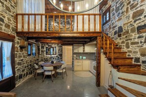 Un intérieur rustique avec un mur en pierre, un escalier en bois, un coin repas avec une table ronde et des chaises, et une cuisine équipée de meubles et d’appareils électroménagers.
