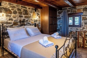 A bedroom with a stone wall, a wooden ceiling, a bed with white linens, and a window with curtains. - Vorenia Horizon (Viannos)
