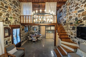 A rustic interior with a stone wall, wooden staircase, a dining area with a round table, and a chandelier. - Vorenia Horizon (Viannos)