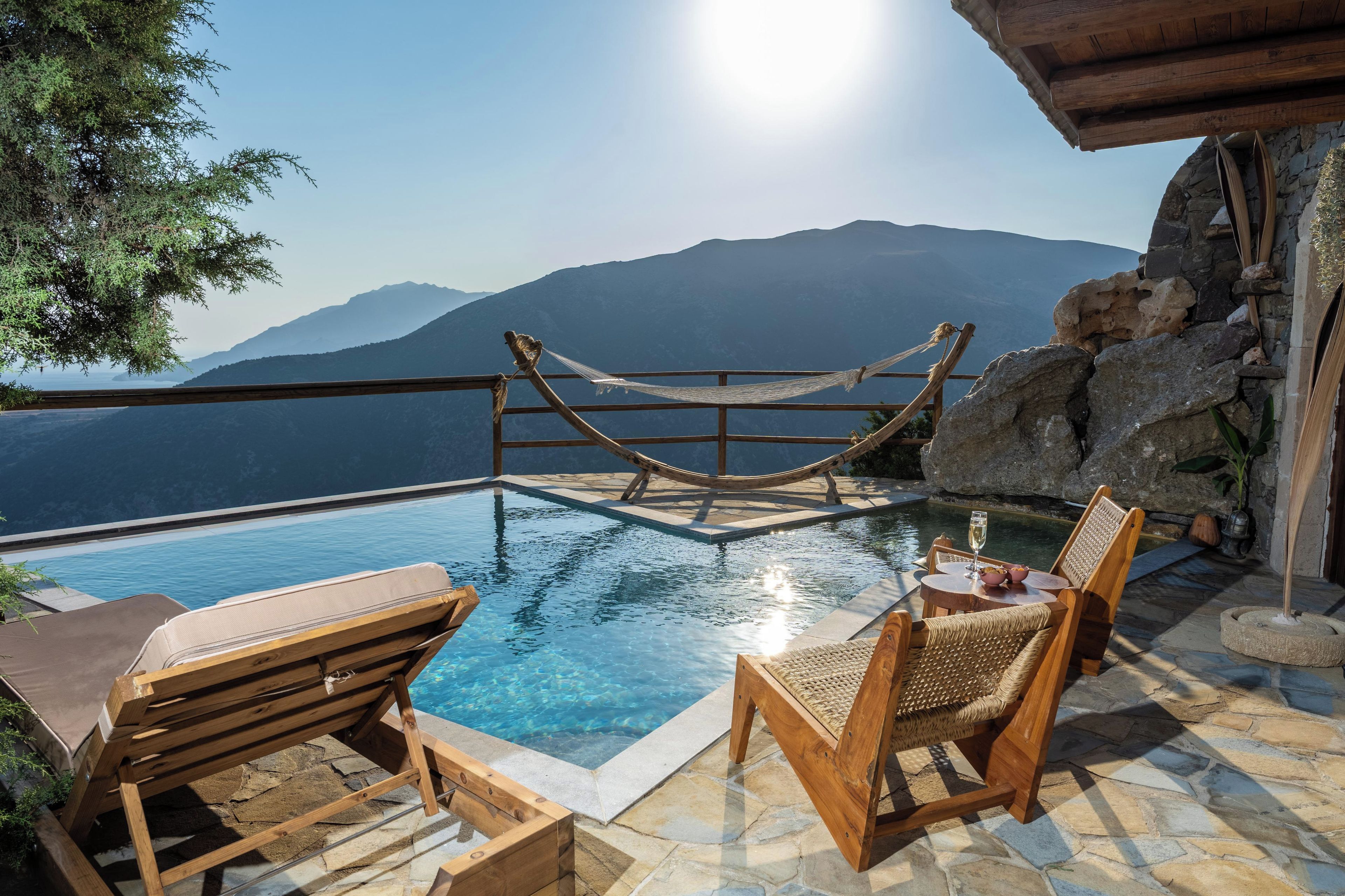 A pool area with wooden loungers and a hammock, overlooking a mountainous landscape.