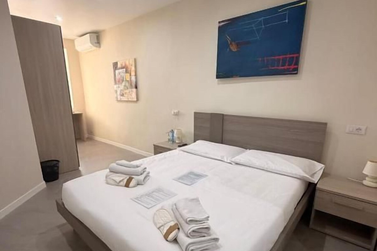 A modern hotel room with a large bed, a bedside table, a nightstand, and a wall-mounted artwork.