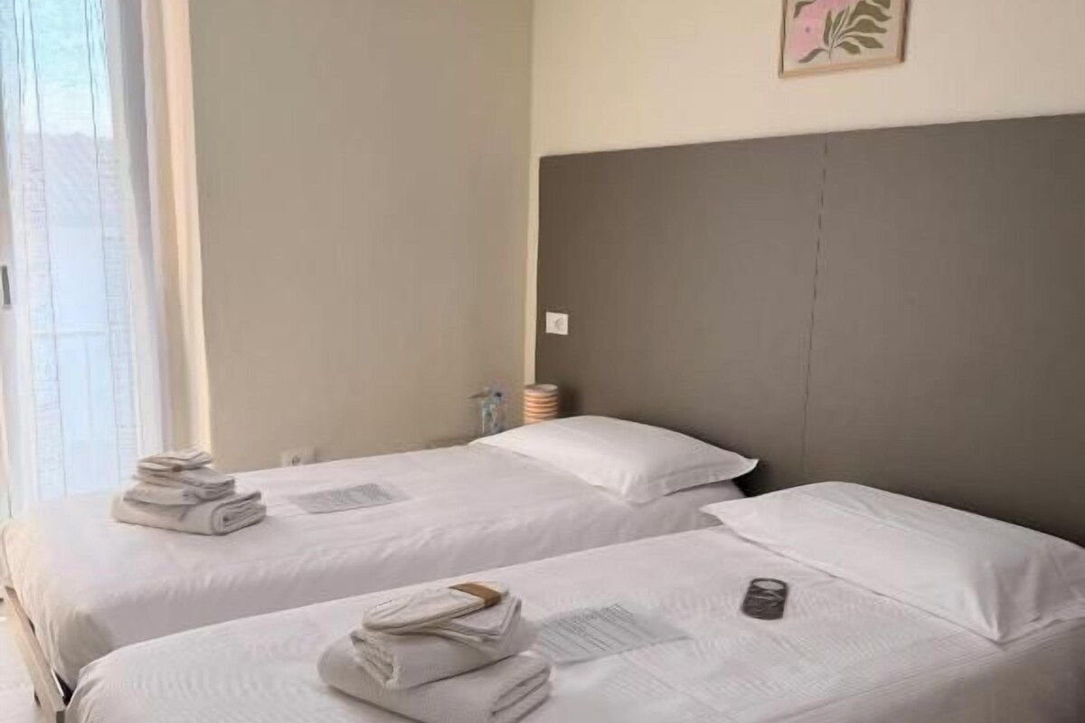 A hotel room with two beds, a large window, an air conditioning unit, and a framed artwork on the wall.