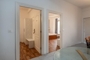 Un pasillo que conduce a un dormitorio con cama y ventana, un baño con ducha, y un comedor con mesa de vidrio.