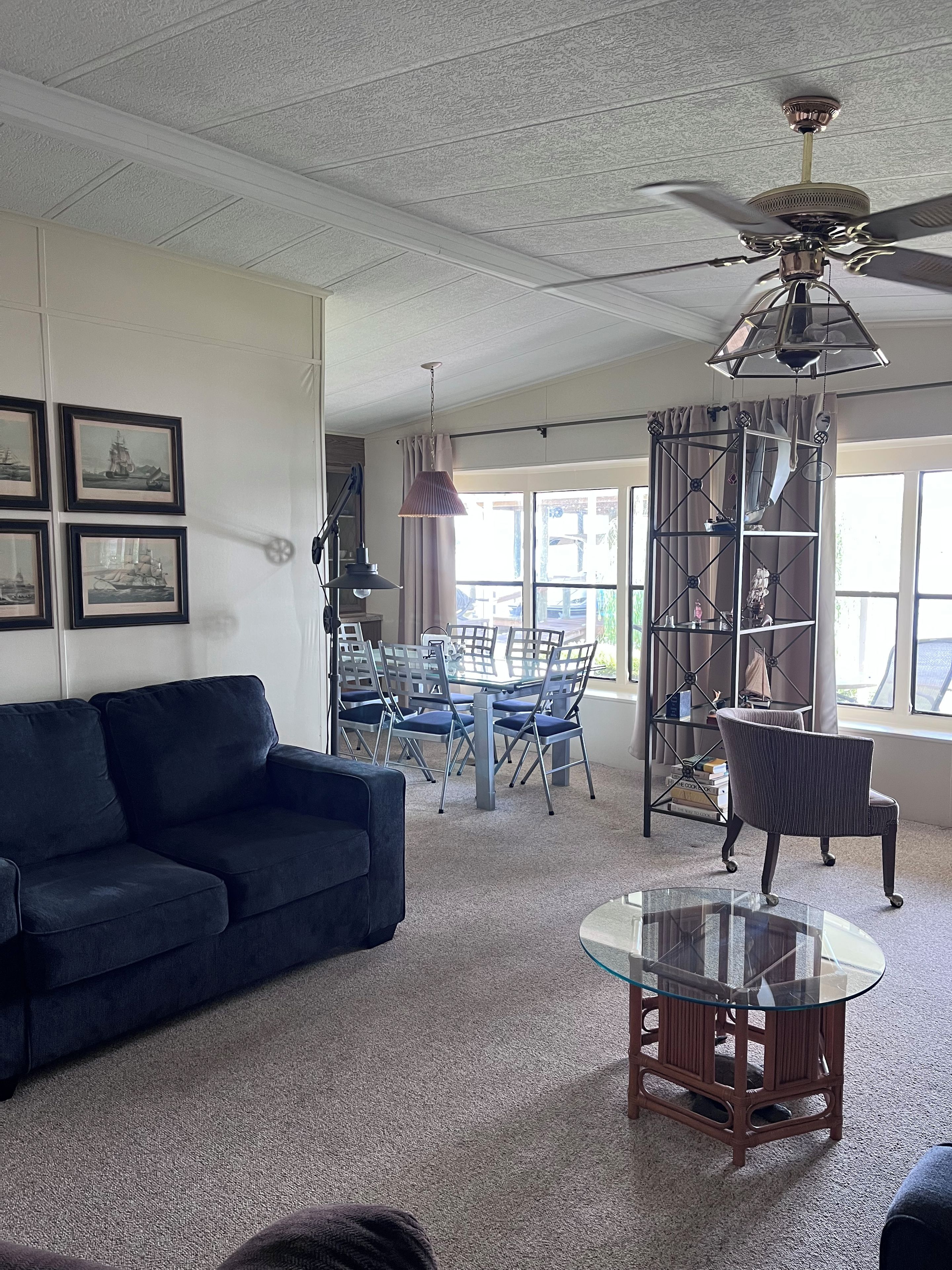 A living room with a dark blue sofa, a glass coffee table, a dining area with a glass table and chairs, and a ceiling fan.