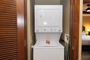 A compact washer and dryer unit with a control panel, located in a hotel room with wooden blinds and a bed visible in the background. - Peakside at One Ski Hill: Peak 8 Ski-In/Out, 2 Baths, A/C, Pools, Shuttle (Breckenridge)