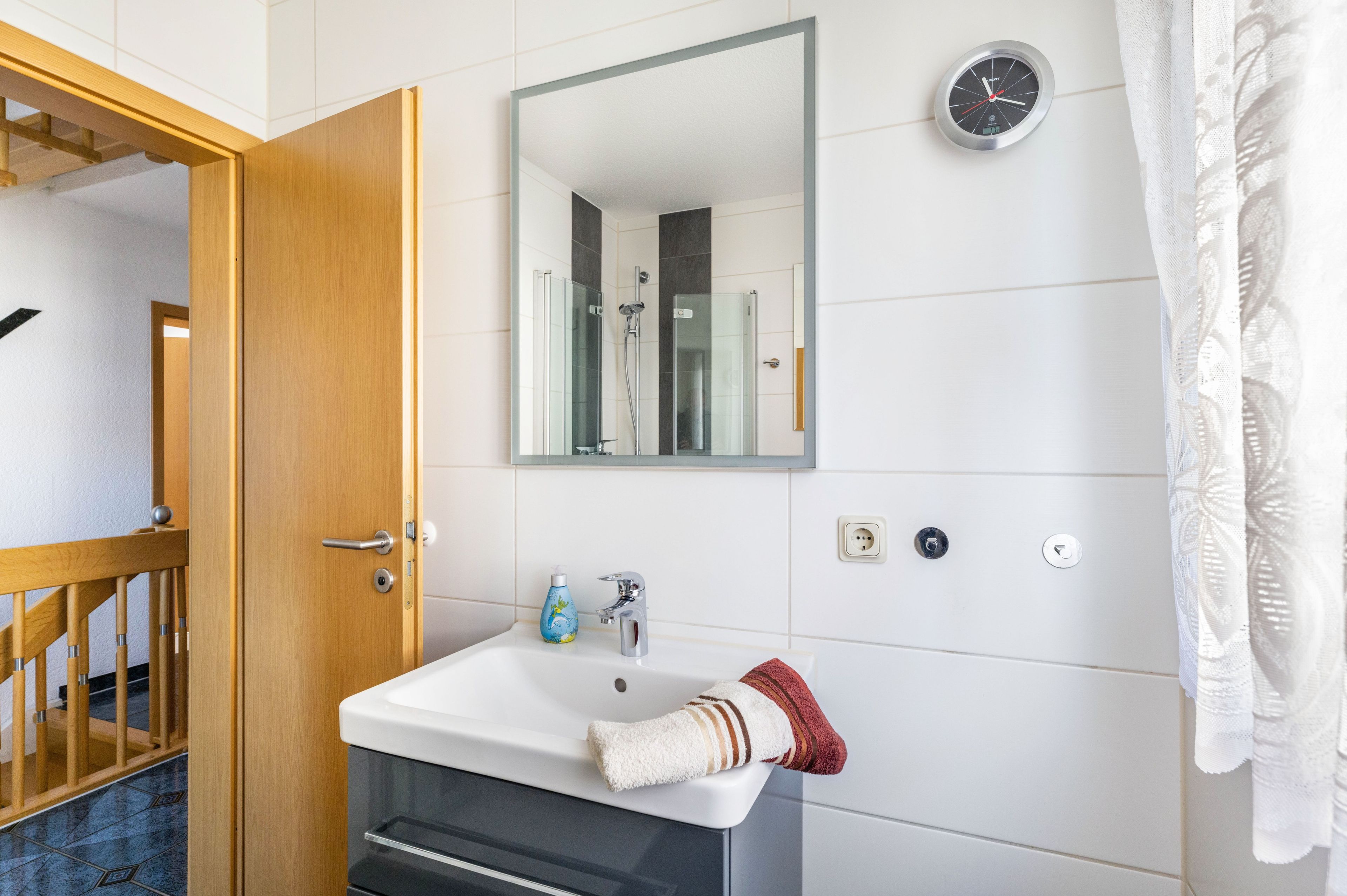 Ein Badezimmer mit Holztür, Waschbecken in Weiß, Spiegel, Uhr und Handtuchhalter.