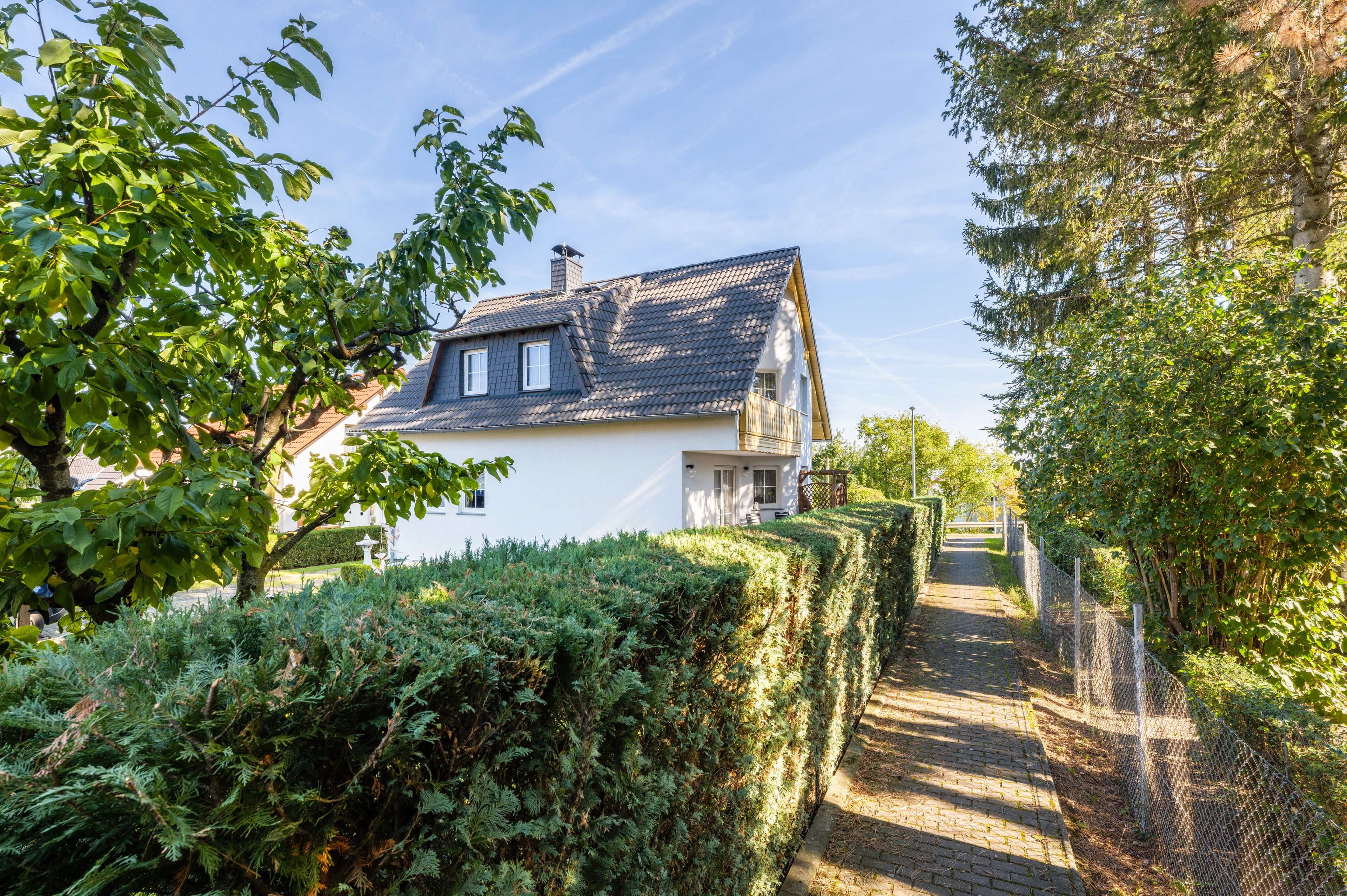 A white house with a sloped roof, surrounded by greenery and a paved path leading to a gate.