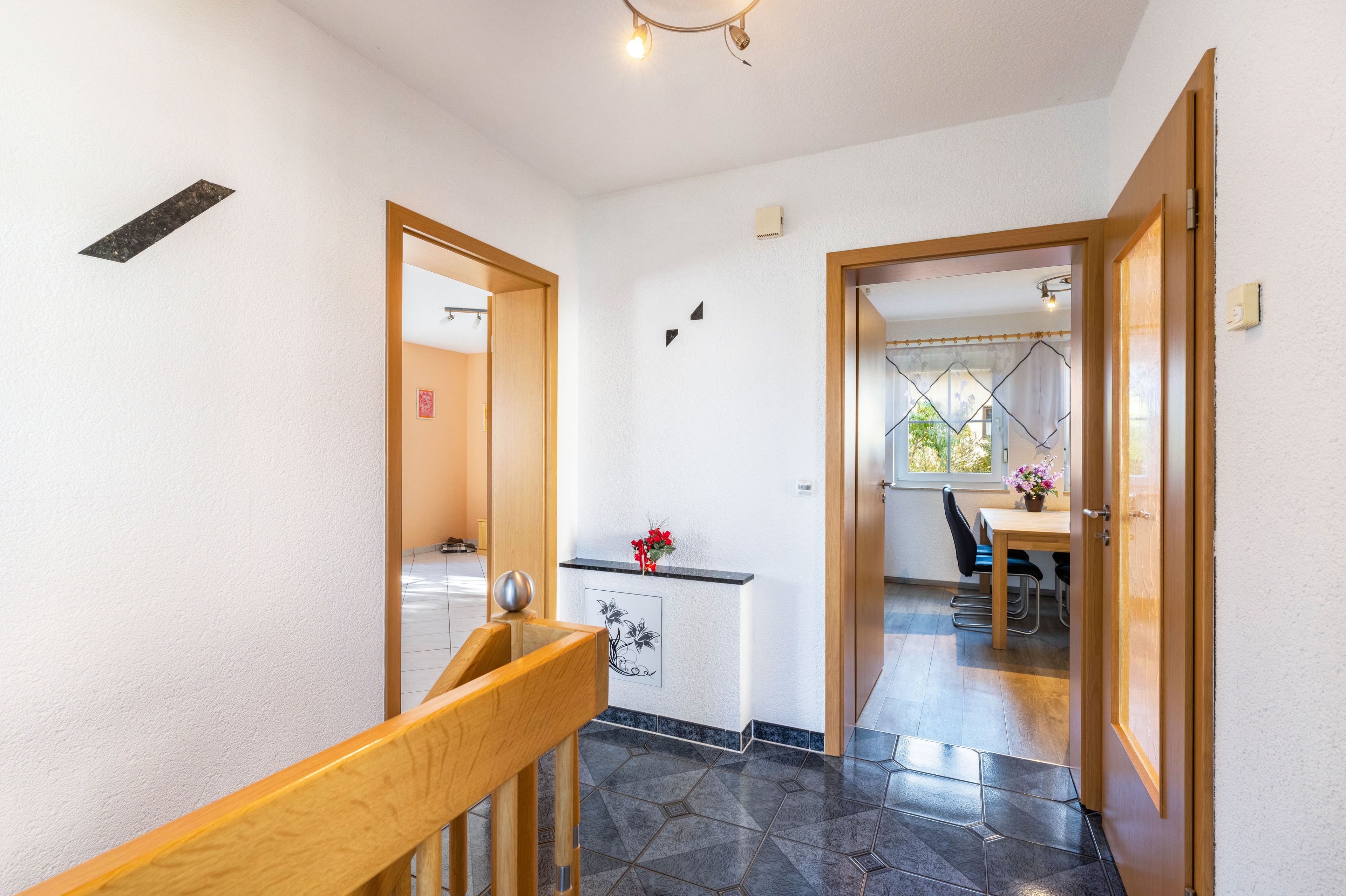 Un pasillo con una escalera de madera, una pequeña cocina con un lavamanos y un jarrón de flores, y un comedor con una mesa y sillas.