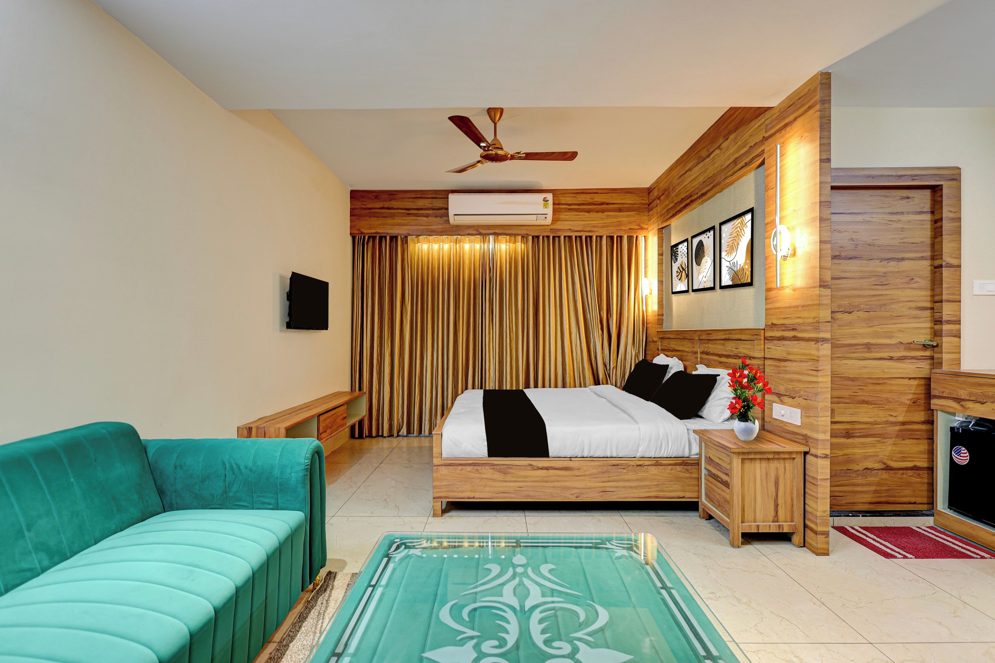 Un dormitorio con cama, cabecera de madera, ventilador de techo, unidad de aire acondicionado, mesita de noche con lámpara, sofá verde y una mesa de vidrio decorativa.