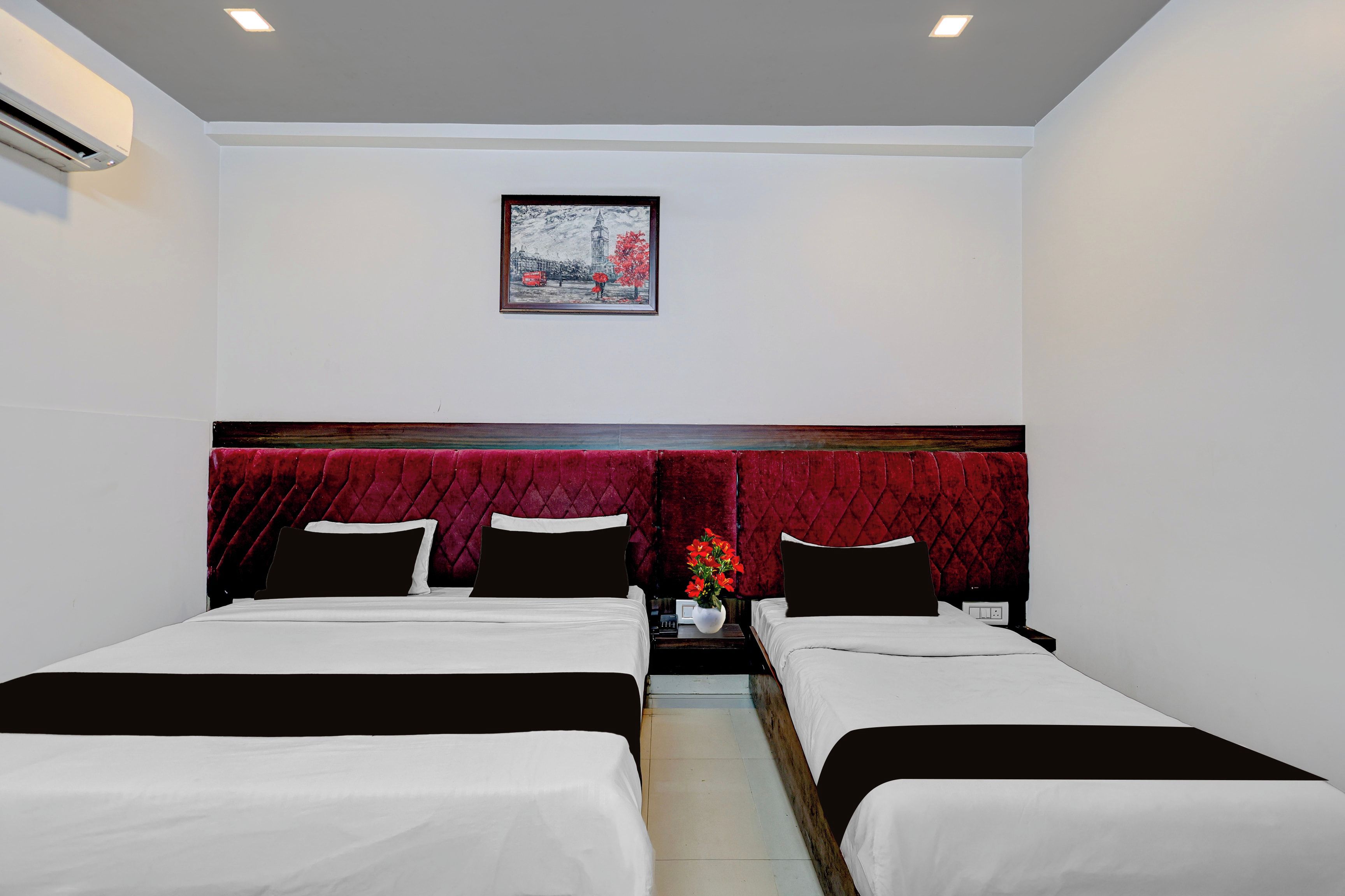 A hotel room with two beds, a red headboard, a framed picture on the wall, and a nightstand with a vase of flowers.