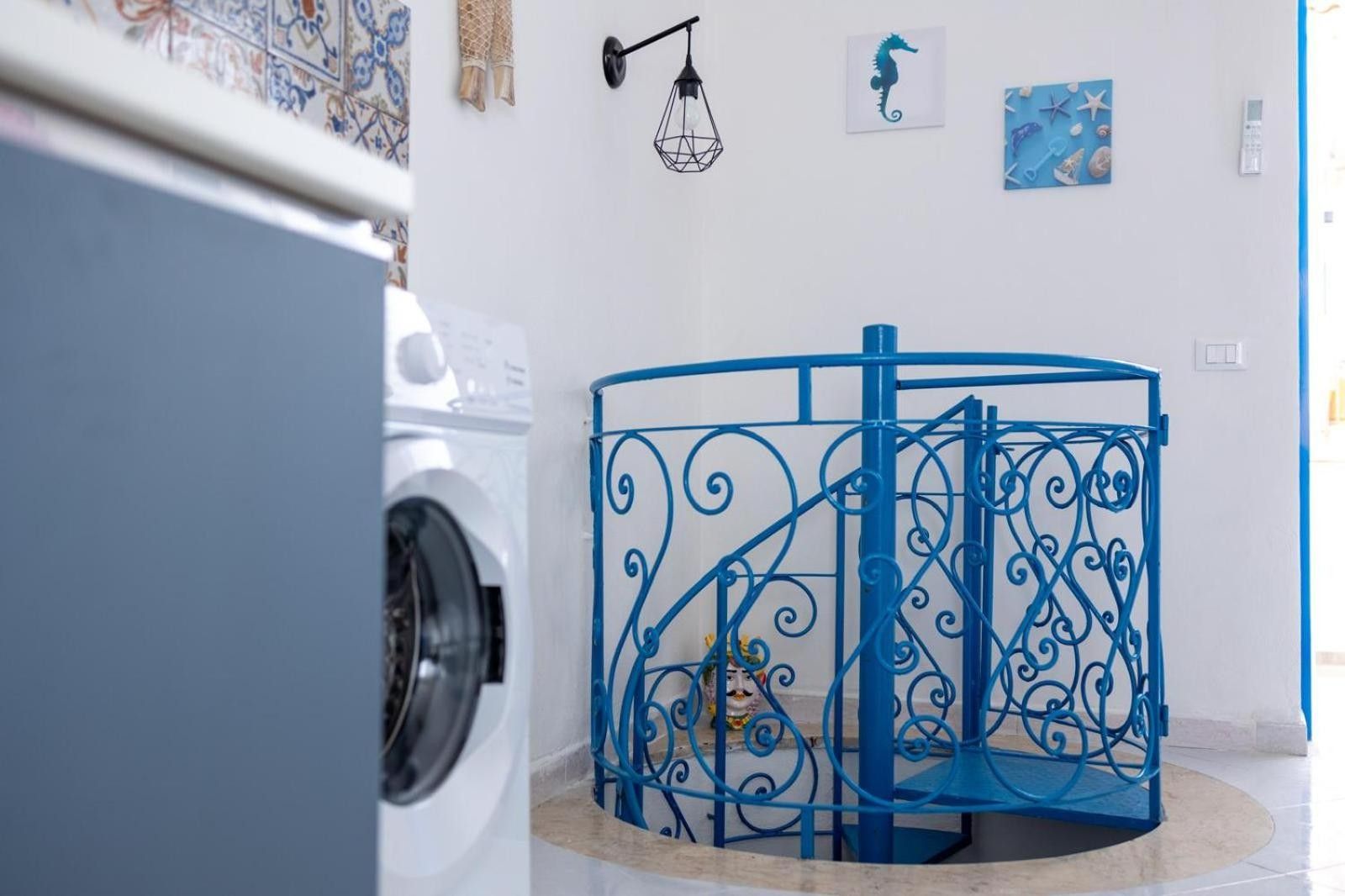 Ein Waschbereich mit einer Waschmaschine, einem blauen Metall-Wäschesammler und einer Wand mit Dekoration, darunter ein Gemälde von einem Seepferdchen und einer geometrischen Leuchte.