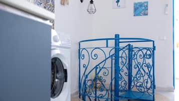 Ein Waschbereich mit einer Waschmaschine, einem blauen Metall-Wäschesammler und einer Wand mit Dekoration, darunter ein Gemälde von einem Seepferdchen und einer geometrischen Leuchte.