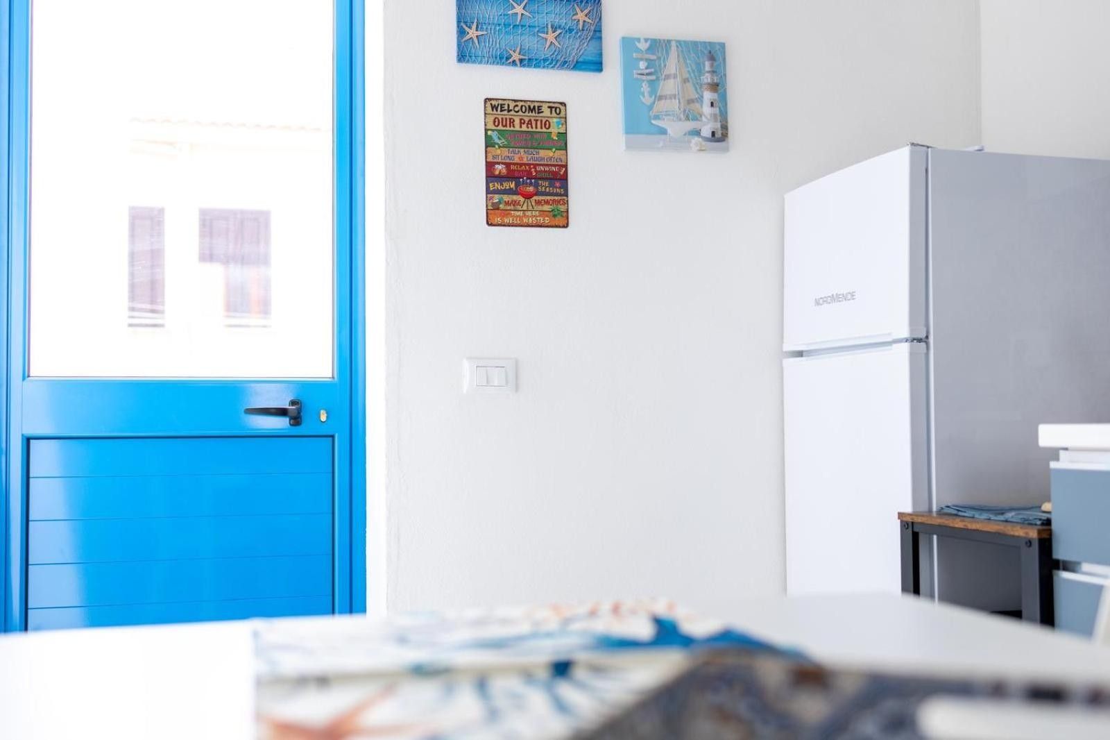 Una stanza con una porta blu, un muro bianco con un cartello di benvenuto, un frigorifero e un tavolino con un libro.