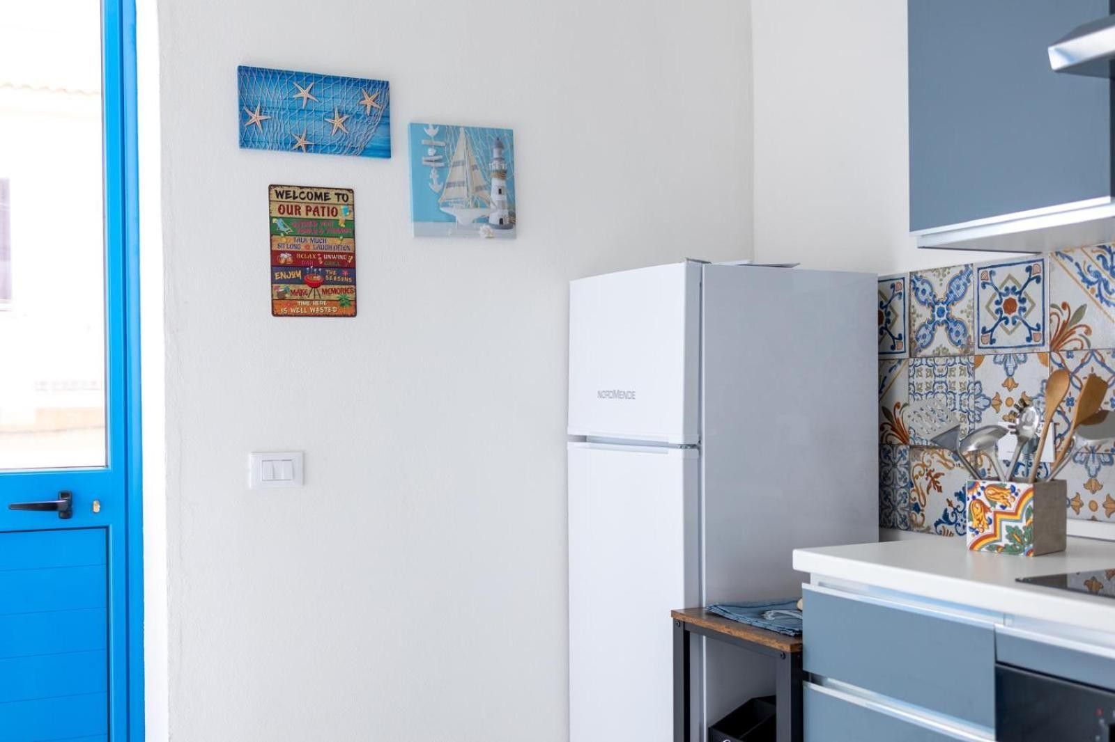 Een keuken met een witte koelkast, een blauwe deur en decoratieve wandkunst, waaronder een vuurtoren en een welkomstbord.