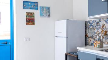 Een keuken met een witte koelkast, een blauwe deur en decoratieve wandkunst, waaronder een vuurtoren en een welkomstbord.
