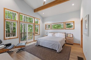 Ein Schlafzimmer mit einem großen Bett, einem Schreibtisch und einem Stuhl. Mehrere Fenster sorgen für natürliches Licht und Blick auf Bäume.